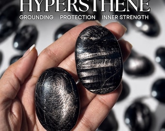 Puede incluir: Dos piedras de hiperstena pulidas de forma ovalada con un brillo metálico oscuro y texturas lineales visibles. El texto HYPERSTHENE, GROUNDING, PROTECTION, INNER STRENGTH aparece sobre las piedras sostenidas frente a un fondo borroso.