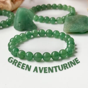 Puede incluir: Pulseras de cuentas de aventurina verde y piedras de aventurina en bruto. Las pulseras est&aacute;n hechas de cuentas verdes redondas y pulidas. El texto "GREEN AVENTURINE" est&aacute; impreso en verde debajo. El fondo es blanco.