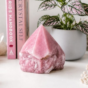 May include: A polished pink rhodonite crystal point with a natural, rough base. The stone features a vibrant rose-colored hue with subtle white and dark mineral inclusions, standing upright as a decorative mineral specimen on a flat surface.