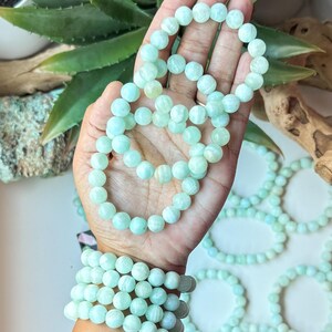 May include: Several strands of light blue-green beaded bracelets. One hand holds a strand of the bracelets, while another arm displays multiple bracelets stacked on the wrist. The beads are round and have a matte finish. The background includes green plants.