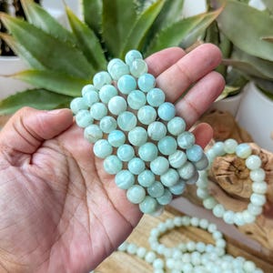 May include: Several strands of light blue gemstone bracelets. The bracelets are made of round, polished beads. The bracelets are held in a hand, with other bracelets and strands of beads visible in the background. The beads have a subtle, natural pattern.