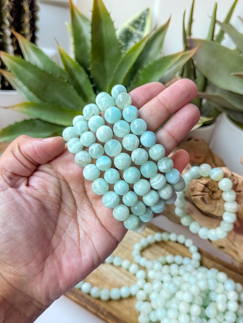 May include: Several strands of light blue gemstone bracelets. The bracelets are made of round, polished beads. The bracelets are held in a hand, with other bracelets and strands of beads visible in the background. The beads have a subtle, natural pattern.
