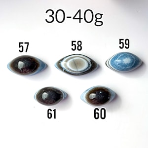 May include: Five oval-shaped stones labelled 57, 58, 59, 60, and 61, weighing 30-40g. Stone 57 is dark brown, 58 has concentric white and brown rings, 59 is blue with white banding, 60 is dark brown, and 61 is dark brown with a smooth finish.