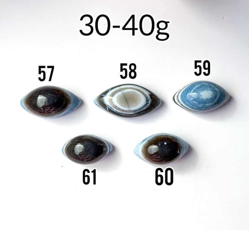 May include: Five oval-shaped stones labelled 57, 58, 59, 60, and 61, weighing 30-40g. Stone 57 is dark brown, 58 has concentric white and brown rings, 59 is blue with white banding, 60 is dark brown, and 61 is dark brown with a smooth finish.