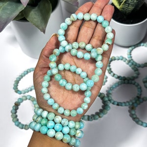May include: Several bracelets made of light blue, round beads are displayed. The bracelets are stacked on a wrist and held in a hand. The beads have a marbled appearance. Other bracelets are scattered in the background.