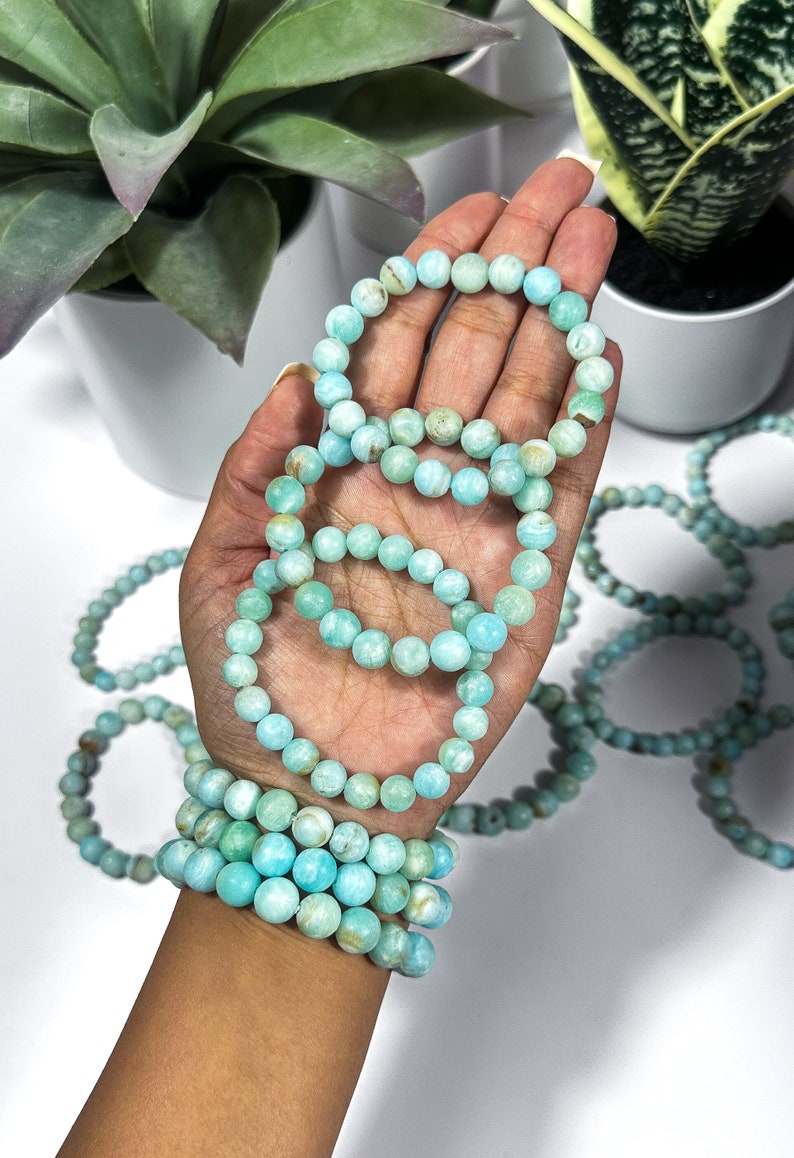 May include: Several bracelets made of light blue, round beads are displayed. The bracelets are stacked on a wrist and held in a hand. The beads have a marbled appearance. Other bracelets are scattered in the background.