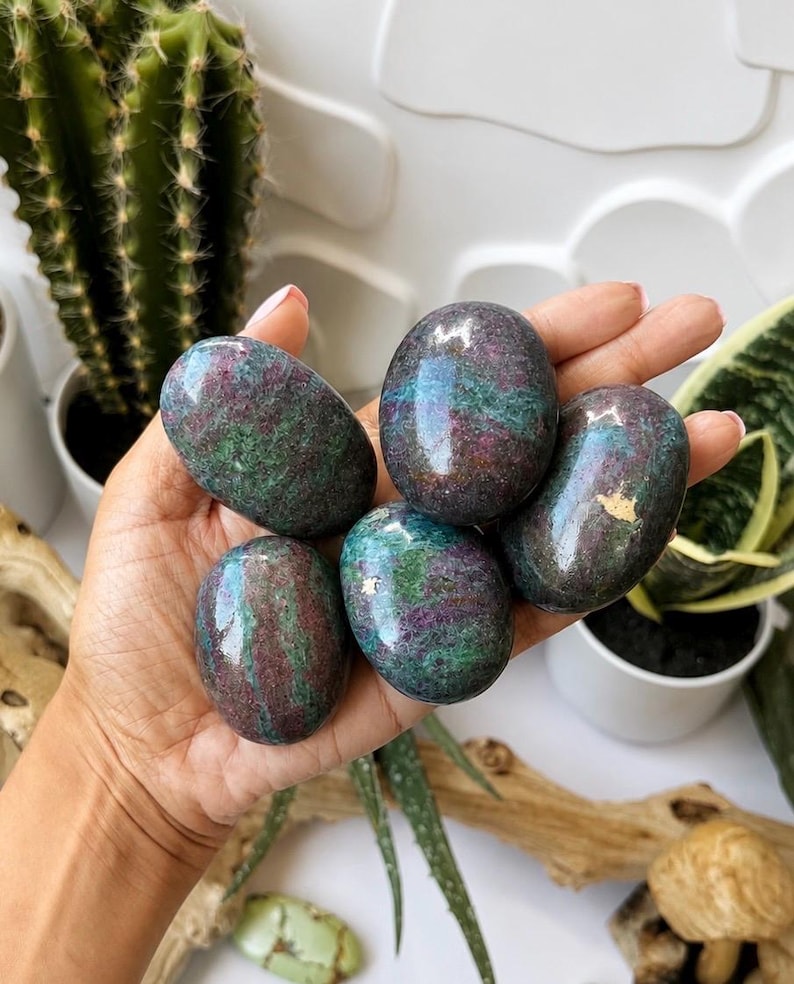 May include: Five polished, oval-shaped ruby in fuchsite stones featuring a mottled blend of deep magenta, dark red, and forest green colours with a smooth, reflective surface. The stones exhibit natural mineral inclusions and variations in texture.
