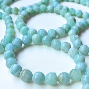 May include: Close-up of several strands of light blue gemstone beads. The beads are round and have a slightly marbled appearance. The beads are strung together, creating a necklace or bracelet. The background is white.