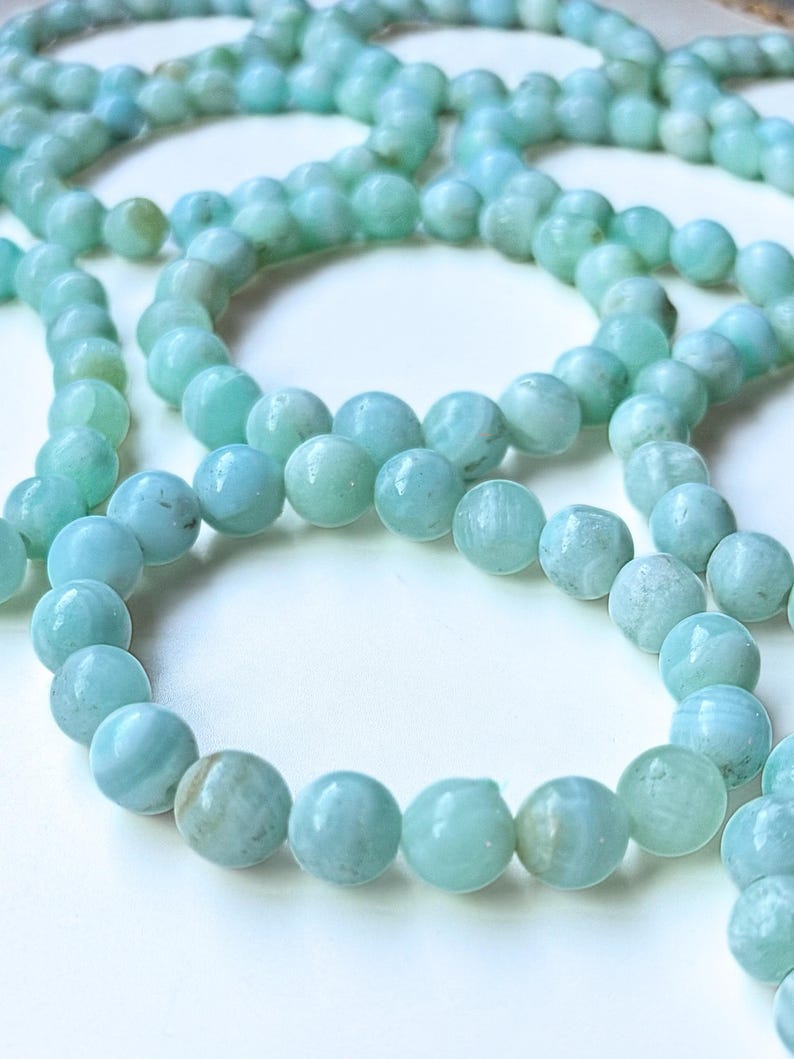 May include: Close-up of several strands of light blue gemstone beads. The beads are round and have a slightly marbled appearance. The beads are strung together, creating a necklace or bracelet. The background is white.