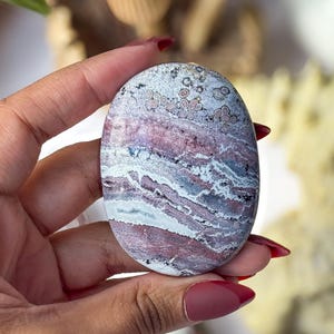 May include: An oval-shaped polished stone featuring a complex pattern of layered bands in shades of dusty rose, deep burgundy, white, and grey, with distinct circular orbicular inclusions visible across the upper portion of the smooth surface.