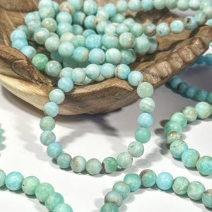 May include: Several strands of light blue and green beaded bracelets. The bracelets are made of round, polished beads. Some bracelets are arranged in a wooden bowl, while others are laid out on a white surface. The beads have a marbled appearance.