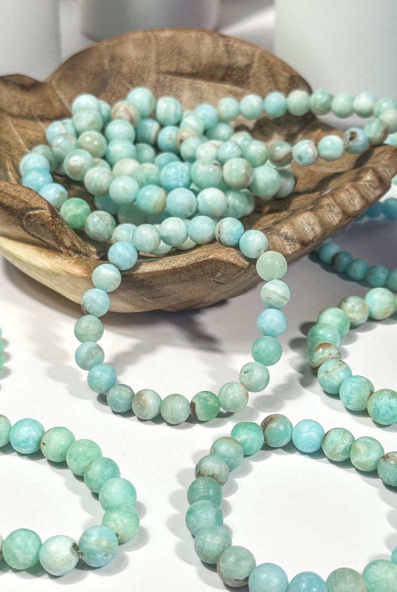 May include: Several strands of light blue and green beaded bracelets. The bracelets are made of round, polished beads. Some bracelets are arranged in a wooden bowl, while others are laid out on a white surface. The beads have a marbled appearance.