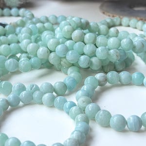 May include: Close-up of several strands of light blue-green gemstone beads. The beads are round and have a slightly translucent appearance. The beads are arranged in a casual, overlapping manner, suggesting they are for jewelry making or crafting.