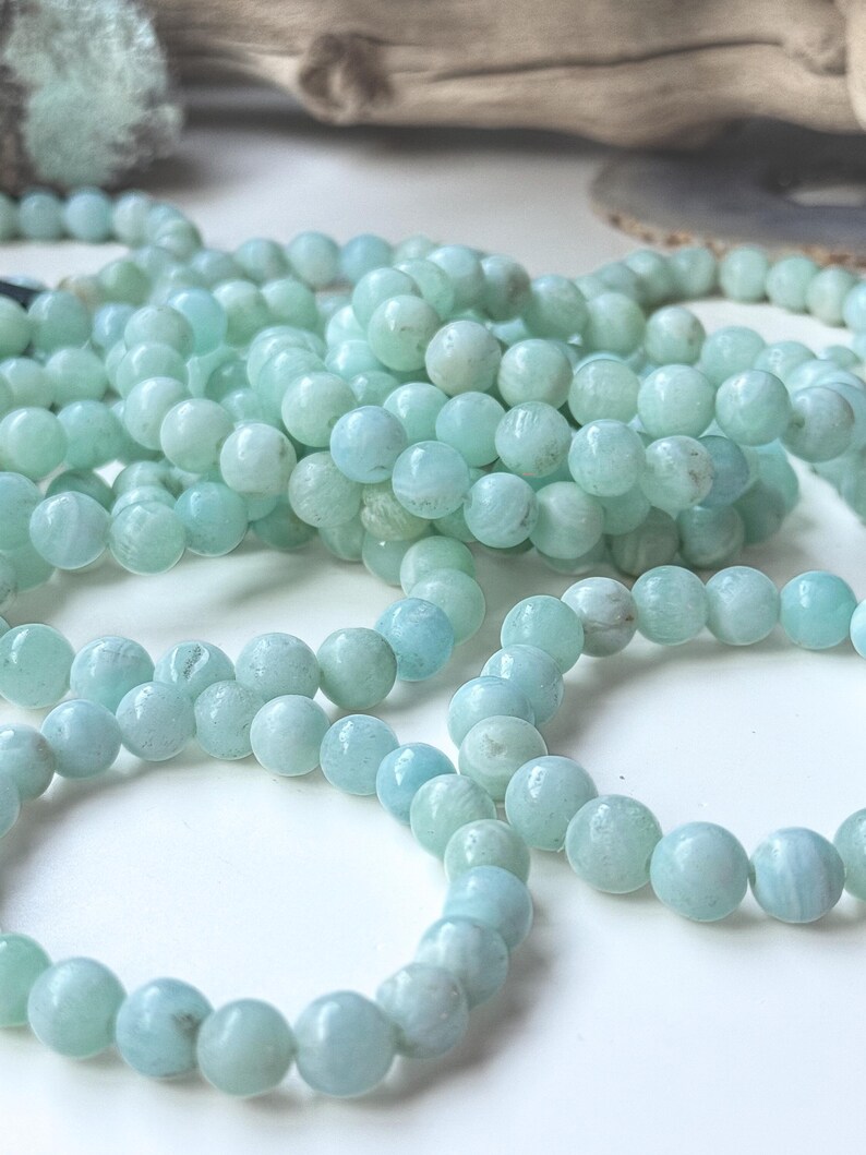 May include: Close-up of several strands of light blue-green gemstone beads. The beads are round and have a slightly translucent appearance. The beads are arranged in a casual, overlapping manner, suggesting they are for jewelry making or crafting.
