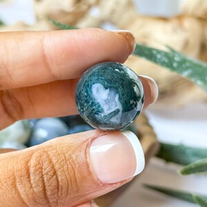 May include: A polished, round gemstone with a swirling pattern of dark green and white. The stone is held between fingers, showcasing its smooth, reflective surface. The gemstone is a natural, decorative item.