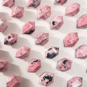 May include: Faceted pink rhodonite crystals with black markings are displayed on a white surface. The crystals' geometric forms and natural colour variations are highlighted. These crystals are suitable for collectors or those interested in gemstones.