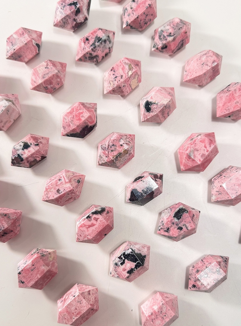 May include: Faceted pink rhodonite crystals with black markings are displayed on a white surface. The crystals' geometric forms and natural colour variations are highlighted. These crystals are suitable for collectors or those interested in gemstones.