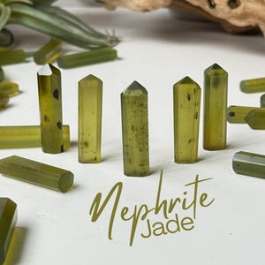 May include: A collection of Nephrite Jade crystals in various shapes and sizes, with a light green hue. The crystals are arranged on a white surface, with the text "Nephrite Jade" in a cursive font.