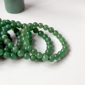 Puede incluir: Varias hebras de pulseras de cuentas de piedra aventurina verde. Las cuentas son redondas y pulidas. Un objeto cil&iacute;ndrico verde est&aacute; en el fondo. Las pulseras se exhiben sobre una superficie blanca.