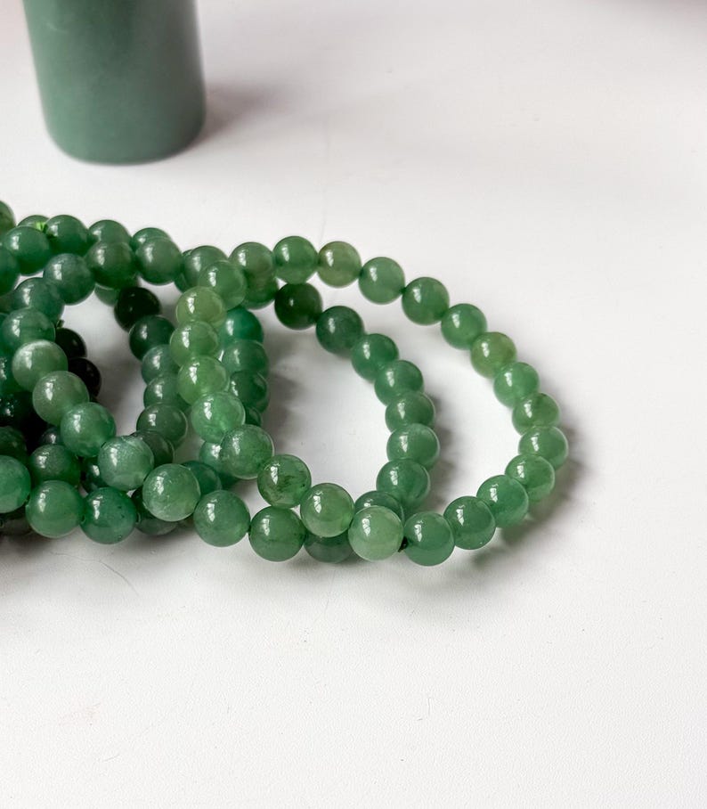 Puede incluir: Varias hebras de pulseras de cuentas de piedra aventurina verde. Las cuentas son redondas y pulidas. Un objeto cil&iacute;ndrico verde est&aacute; en el fondo. Las pulseras se exhiben sobre una superficie blanca.