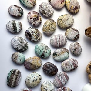 May include: A collection of smooth, oval-shaped polished stones featuring a diverse array of natural patterns and colours, including shades of green, red, brown, white, and yellow, with speckled, marbled, and banded textures throughout the surfaces.