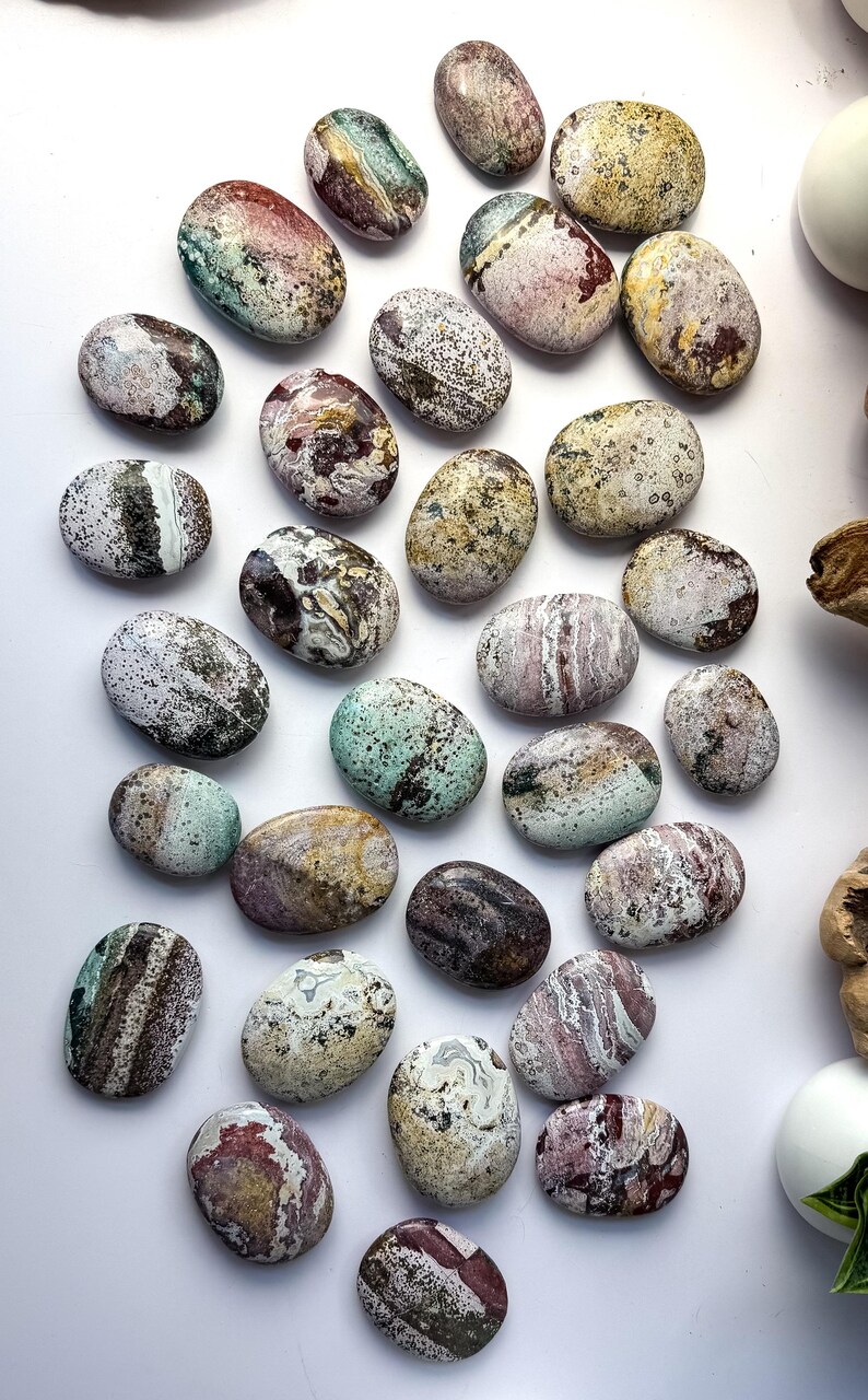 May include: A collection of smooth, oval-shaped polished stones featuring a diverse array of natural patterns and colours, including shades of green, red, brown, white, and yellow, with speckled, marbled, and banded textures throughout the surfaces.