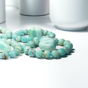 May include: Close-up of Amazonite gemstone bracelets and stones. The bracelets feature round, light blue-green beads with natural variations. The stones are a similar color, with a mix of rounded and rectangular shapes. The background is white.