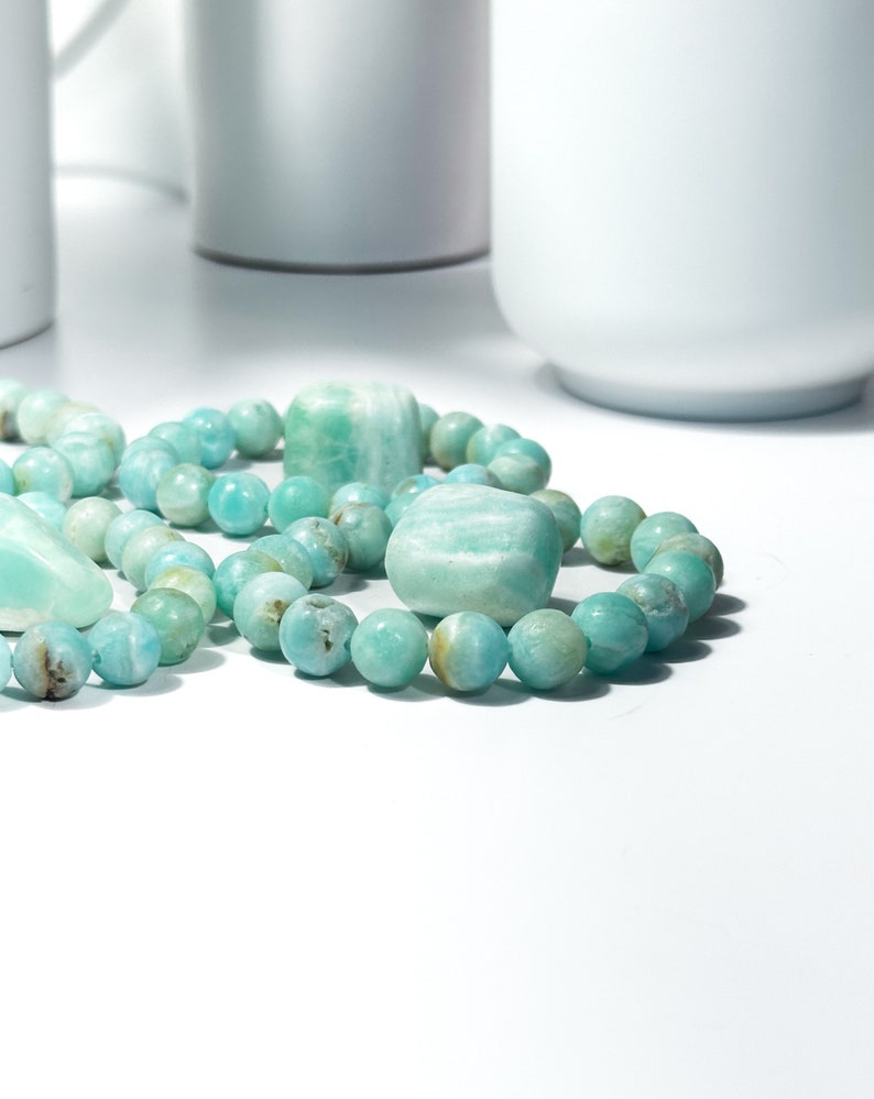 May include: Close-up of Amazonite gemstone bracelets and stones. The bracelets feature round, light blue-green beads with natural variations. The stones are a similar color, with a mix of rounded and rectangular shapes. The background is white.