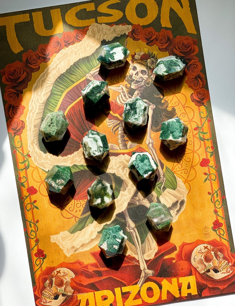 May include: Several faceted green and white moss agate crystals are displayed on a vintage poster. The poster has the word "TUCSON" at the top and a Day of the Dead illustration with roses and a skeleton.