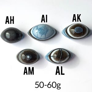 May include: Five oval-shaped stones labelled AH, AI, AK, AM, and AL. AH is dark grey, AI is light blue with white patterns, AK has brown and blue rings, AM is dark brown with a white centre, and AL is brown with blue edges. Text reads 50-60g.