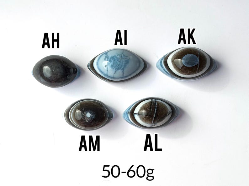 May include: Five oval-shaped stones labelled AH, AI, AK, AM, and AL. AH is dark grey, AI is light blue with white patterns, AK has brown and blue rings, AM is dark brown with a white centre, and AL is brown with blue edges. Text reads 50-60g.