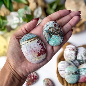 May include: Two polished, oval-shaped stones with intricate patterns of teal, white, red, and brown. The larger stone features a distinct horizontal band of dark brown and yellow, while the smaller stone displays a mottled, speckled texture.