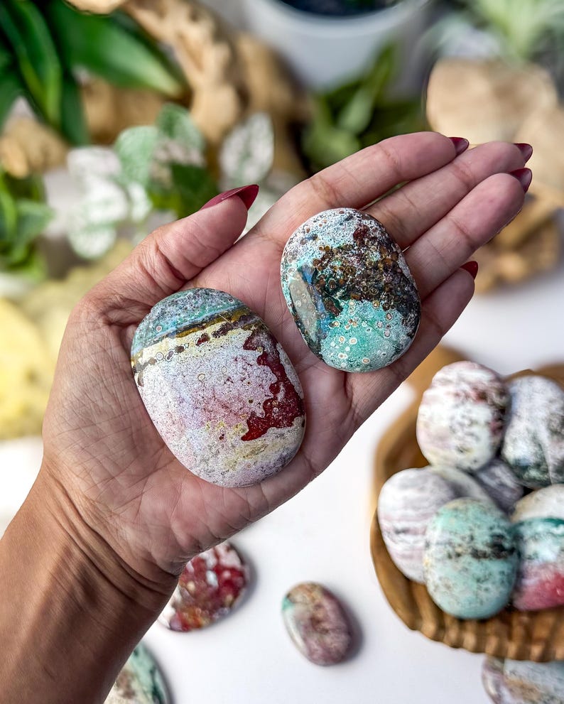 May include: Two polished, oval-shaped stones with intricate patterns of teal, white, red, and brown. The larger stone features a distinct horizontal band of dark brown and yellow, while the smaller stone displays a mottled, speckled texture.