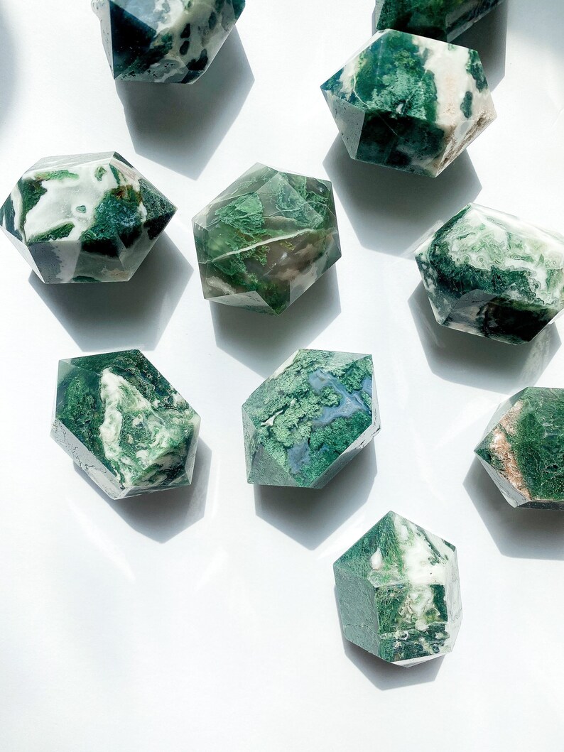 May include: A selection of hexagonal-shaped moss agate crystals. The crystals feature a mix of translucent white and opaque green patterns. The crystals are arranged on a white surface, with shadows adding depth to the composition.