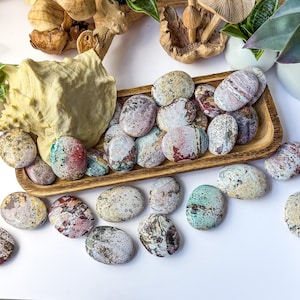 May include: A collection of smooth, oval-shaped polished stones featuring a variety of speckled and marbled patterns in shades of pink, green, cream, and grey. The stones are arranged both inside and around a rectangular wooden tray.
