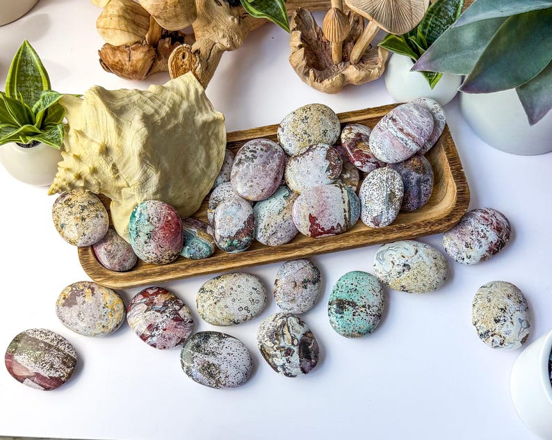 May include: A collection of smooth, oval-shaped polished stones featuring a variety of speckled and marbled patterns in shades of pink, green, cream, and grey. The stones are arranged both inside and around a rectangular wooden tray.