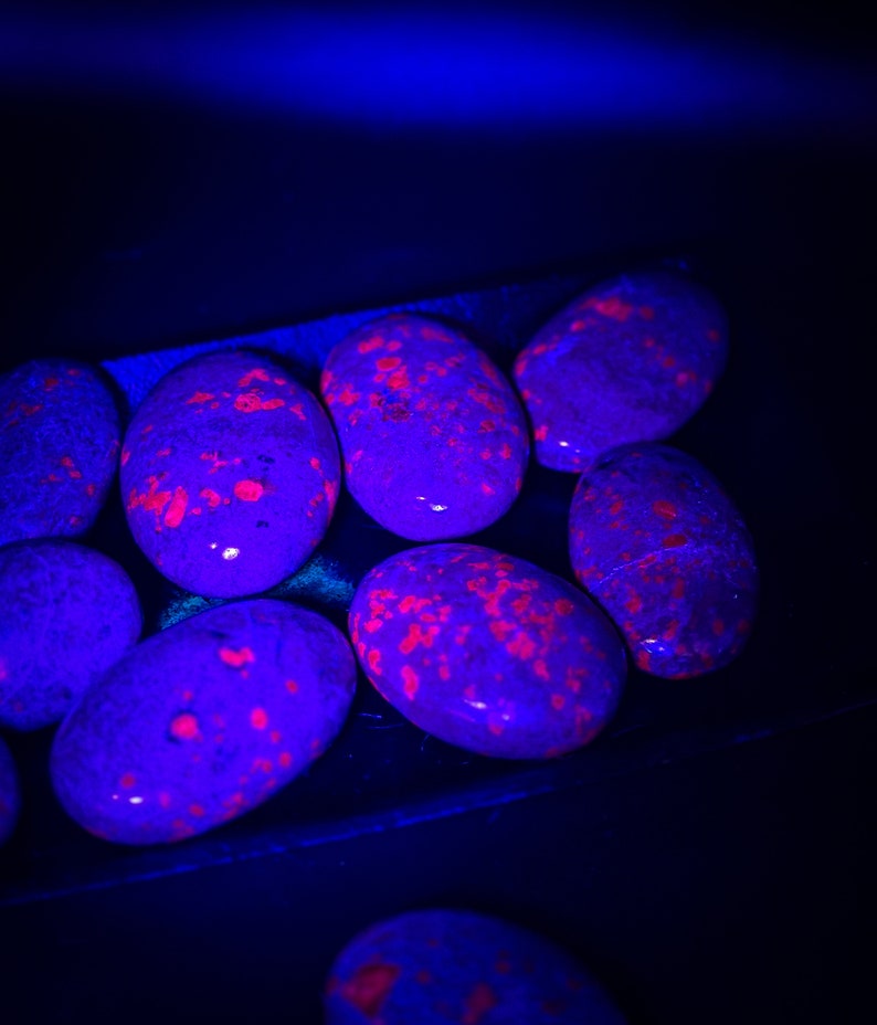 May include: A collection of smooth, oval-shaped stones illuminated under ultraviolet light. The stones appear dark purple and black with vibrant, glowing neon-red or bright orange speckled patterns across their polished surfaces.