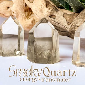 May include: Three smoky quartz crystal points with a light brown hue are displayed on a white surface. The crystals are faceted and have a geometric shape. The text "Smoky Quartz energy transmuter" is written in a decorative font.