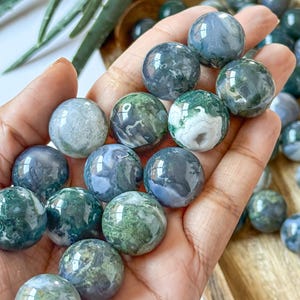 May include: A collection of polished moss agate spheres, each displaying unique patterns of green, white, and blue. The spheres are held in a hand, with additional spheres visible in the background, suggesting a natural stone product.