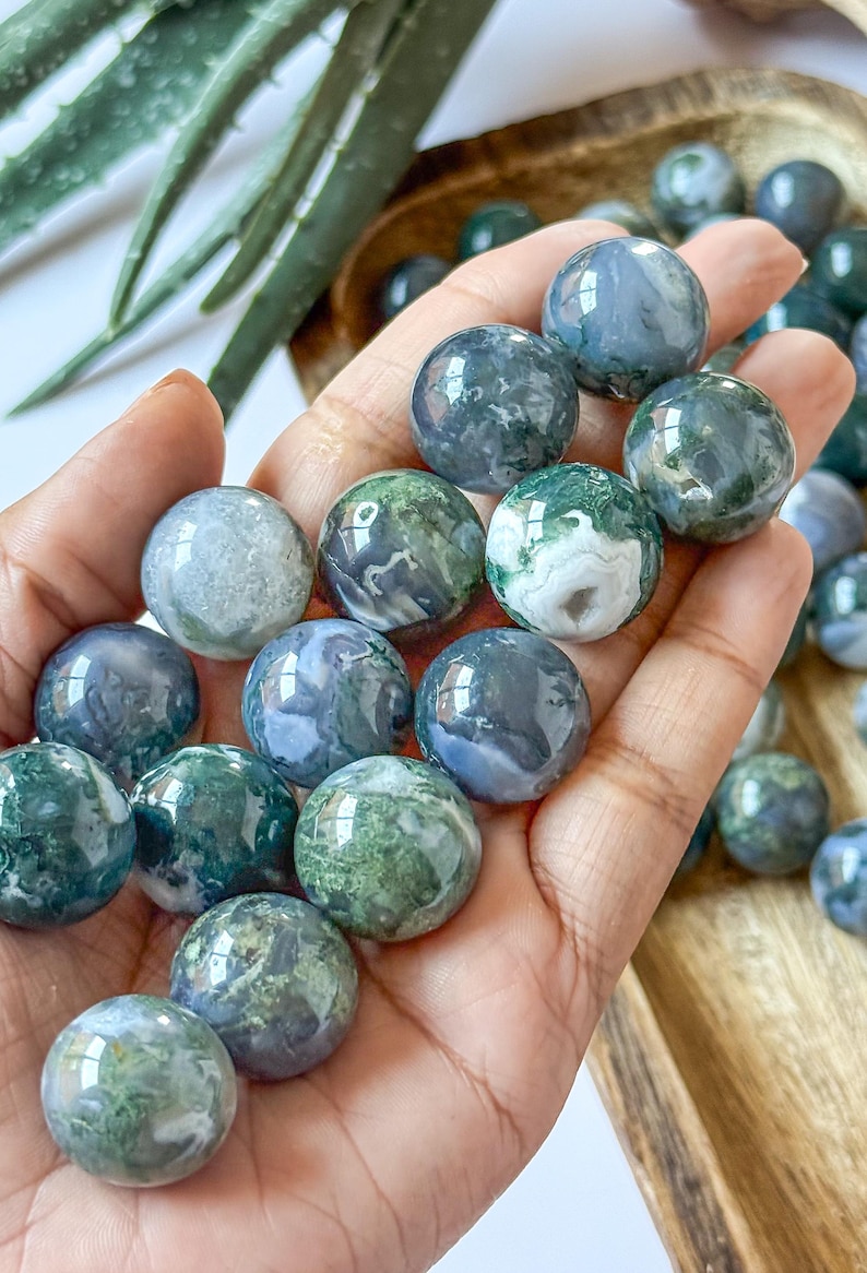 May include: A collection of polished moss agate spheres, each displaying unique patterns of green, white, and blue. The spheres are held in a hand, with additional spheres visible in the background, suggesting a natural stone product.