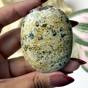 May include: An oval-shaped polished stone featuring a mottled pattern of mustard yellow, forest green, and cream colours. The surface is speckled with small dark green dots and circular inclusions with reddish-brown centres, creating a natural texture.