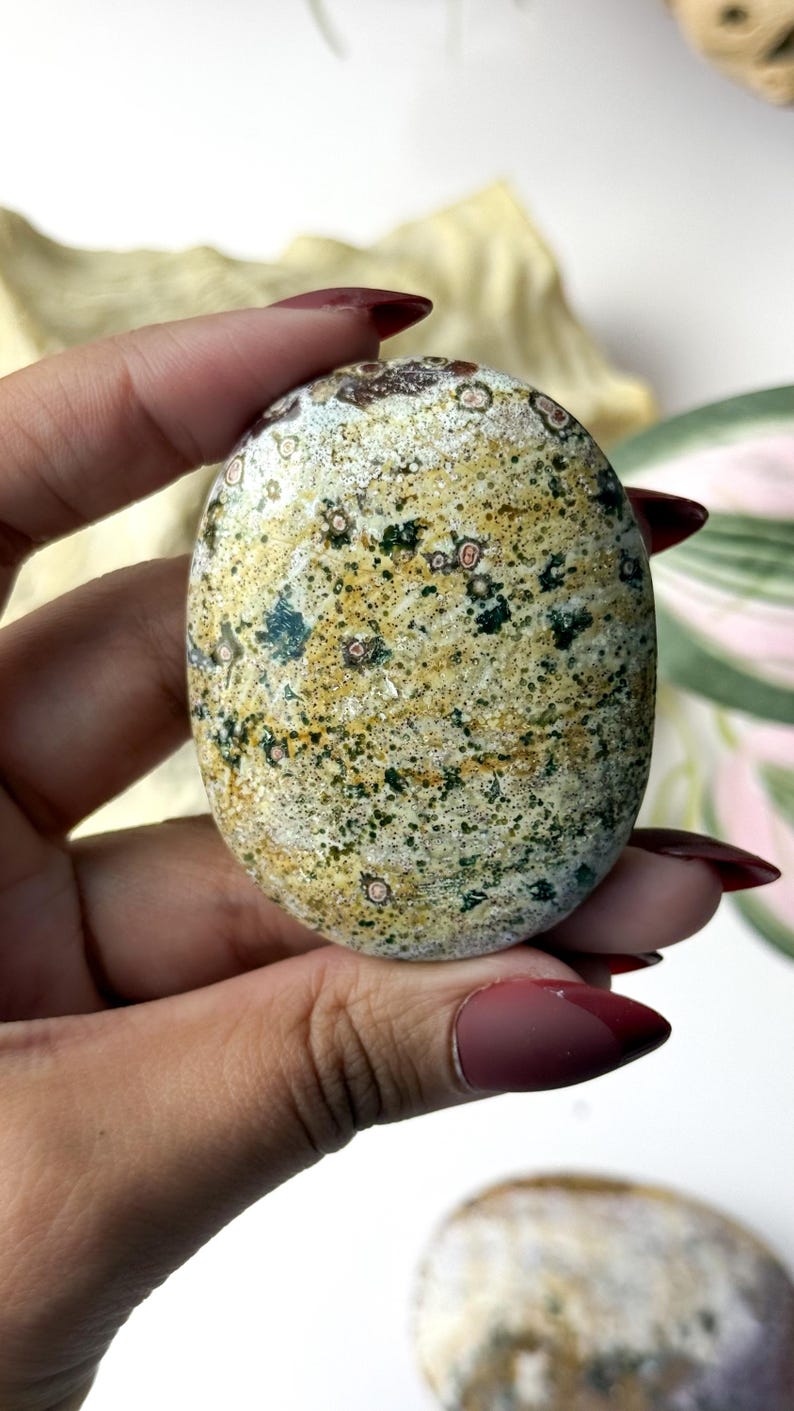 May include: An oval-shaped polished stone featuring a mottled pattern of mustard yellow, forest green, and cream colours. The surface is speckled with small dark green dots and circular inclusions with reddish-brown centres, creating a natural texture.