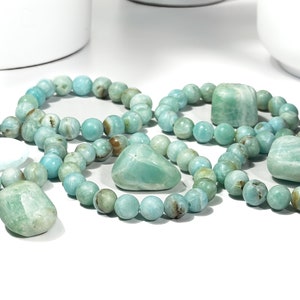May include: Several bracelets and stones made of Amazonite gemstones. The bracelets are composed of round, light blue-green beads. The stones are various shapes and sizes, with a similar color palette. The items are arranged on a white surface.