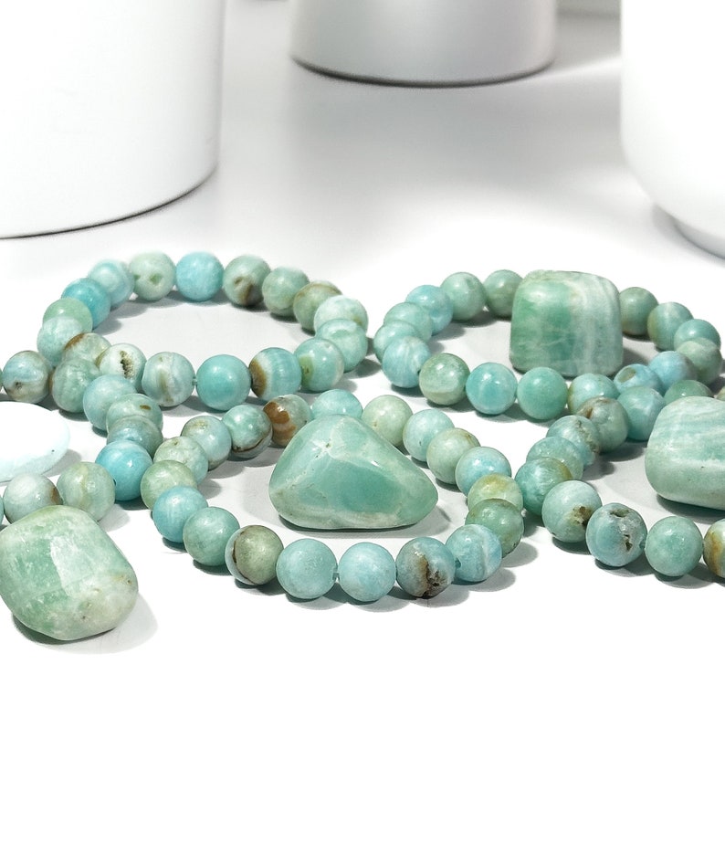 May include: Several bracelets and stones made of Amazonite gemstones. The bracelets are composed of round, light blue-green beads. The stones are various shapes and sizes, with a similar color palette. The items are arranged on a white surface.