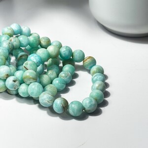 May include: A close-up shot of several strands of turquoise-colored beaded bracelets. The beads are round and have a marbled appearance with variations in color. The bracelets are resting on a white surface, with a blurred background.