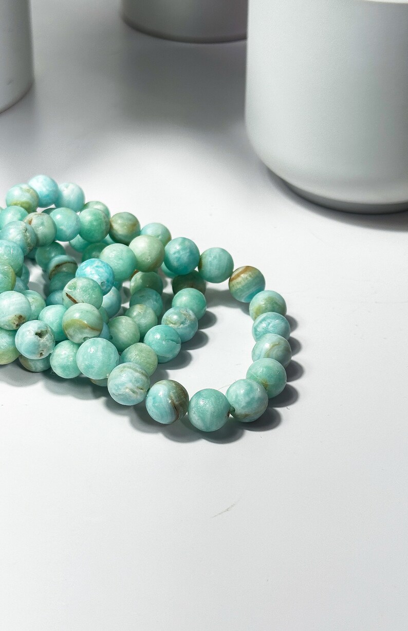 May include: A close-up shot of several strands of turquoise-colored beaded bracelets. The beads are round and have a marbled appearance with variations in color. The bracelets are resting on a white surface, with a blurred background.
