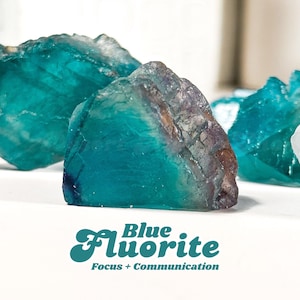 May include: Close-up of several blue fluorite crystals. The crystals have a translucent, glassy appearance with shades of teal and purple. Text at the bottom reads "Blue Fluorite Focus + Communication."
