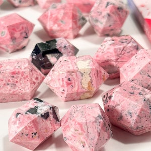 May include: Geometric gemstone crystals in shades of pink and black. The crystals have a marbled pattern and are arranged on a white surface. The crystals are faceted.