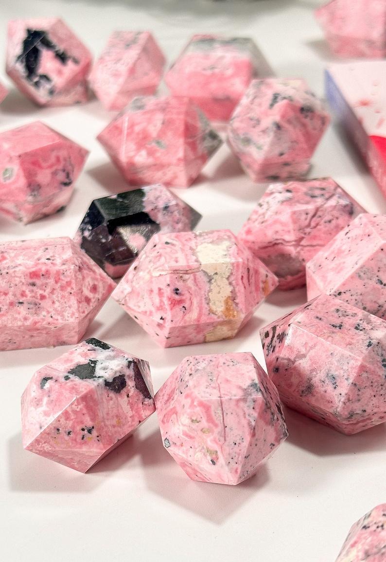 May include: Geometric gemstone crystals in shades of pink and black. The crystals have a marbled pattern and are arranged on a white surface. The crystals are faceted.