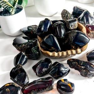 Puede incluir: Una colecci&oacute;n de piedras de &aacute;mbar oscuro, algunas con un tono rojizo, en varias formas y tama&ntilde;os. Algunas piedras est&aacute;n en un cuenco de madera, mientras que otras est&aacute;n esparcidas sobre una superficie blanca.